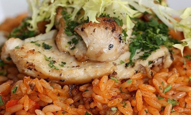 An image of seared chicken on red colored rice, with salad leaves and cilantro on top of it.