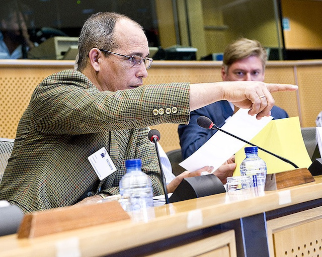 A man in glasses and a checked blazer speaks into a microphone in a conference room, pointing at something.