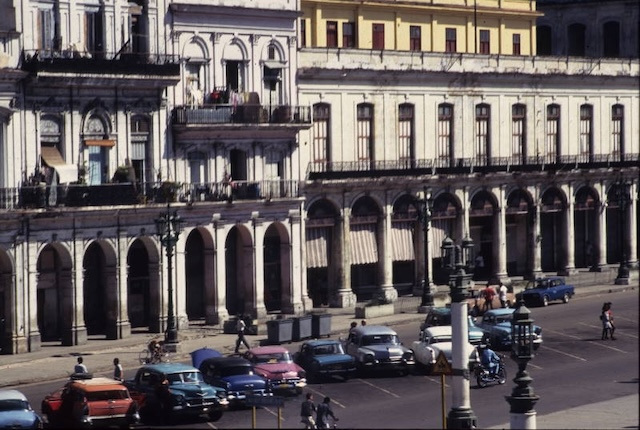 A row of ornate buildings and a parking lot of multicolored antique cars on a a street.