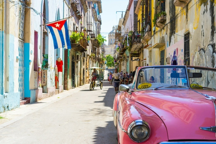 A pink convertible drives down a street with colorful buildings. Pedestrians and a bike taxi follow the car, and a Cuban flag hangs from a doorway. 