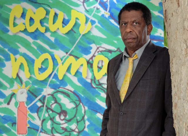 Dany Laferrière, wearing a suit and yellow tie, stands in front of one of his illustrations.