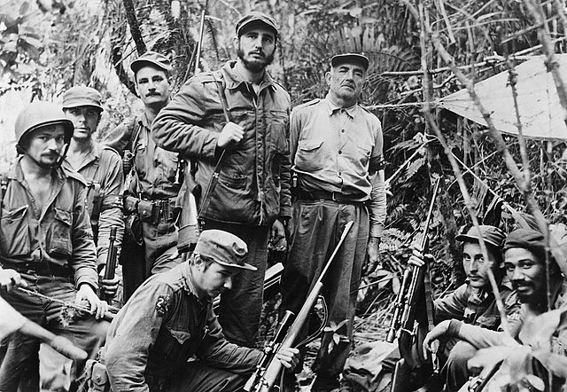 Fidel Castro stands in a forest surrounded by seven men in military uniforms, holding guns.