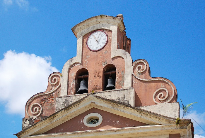 A fading orange-red clock tower with decorative white trim against a blue sky with clouds. The tower has one white clock at the top and two black bells underneath it. 