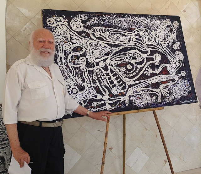 A man with a white beard stands smiling next to a painting of swirling, white skeletal figures against a black-red background.