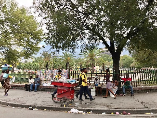 Pedestrians and a vendor's cart on a sidewalk in front of a park with palm trees.