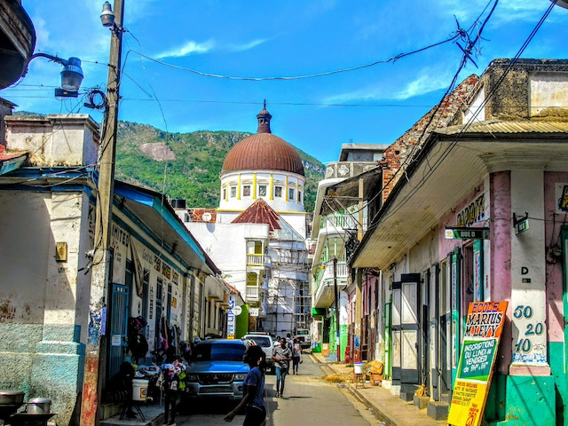 A colorful street in Haiti with several storefronts, parked cars, and a large white church with a brown dome at the end of the block.