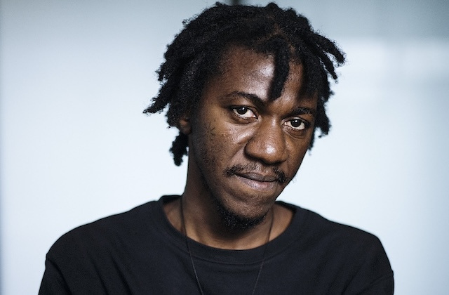 Portrait of a man with dreadlocks in a black sweater against a blue-grey background.