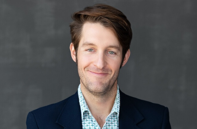 A brown-haired man in a navy suit jacket and light blue patterned shirt against a grey background.