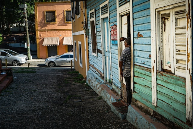 The back of a man standing in a doorway of a blue house with a "se vende"/"for sale" sign, looking out onto the gravel road and paved street with parked cars.