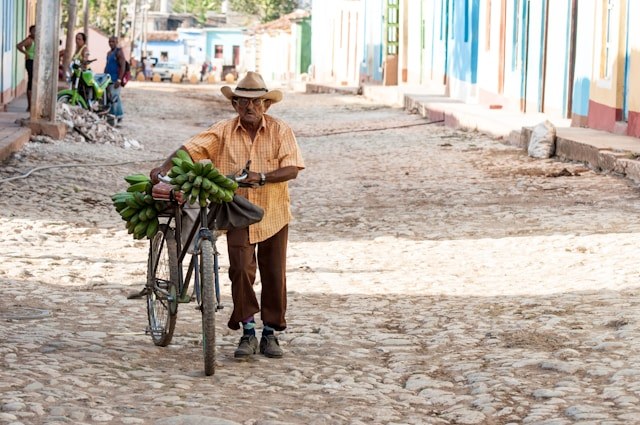 A man in an orange shirt and straw hat walks a bike loaded with plantains along a cobblestone street.