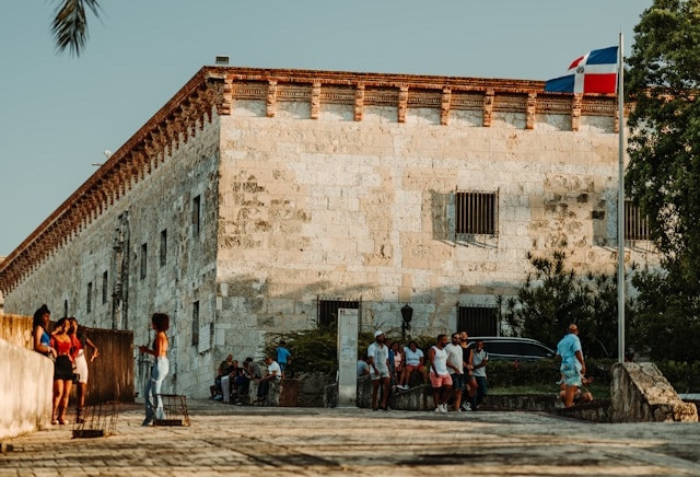 A few groups of people hang out in a town square in front of a large stone building. The Dominican flag flies on a pole.