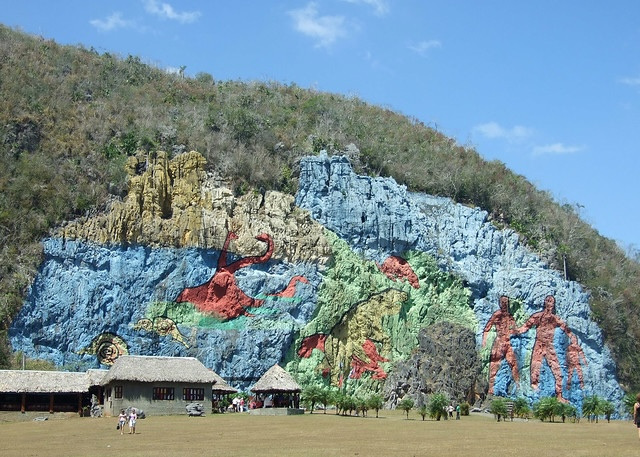 A blue, yellow, green, and red mural of human figures and dinosaurs painted on a giant outcrop of rock.