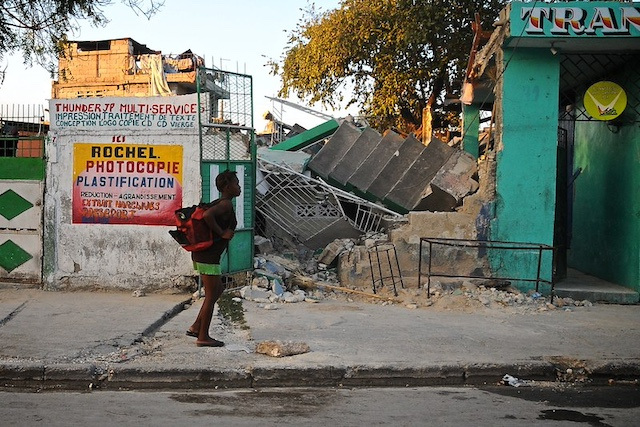 A young Haitian woman in a backpack walks past a demolished building.