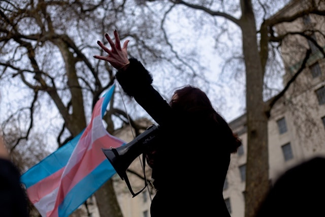 A long-haired person in a dark coat speaks into a bullhorn, arm outstretched, in front of a transgender pride flag.