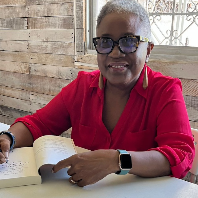 Author Évelyne Trouillot wearing a red shirt, while sitting at a desk, smiling and signing a book.