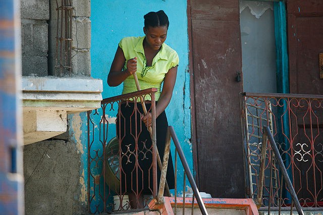 Woman sweeping the porch of a house with a blue wall and an open door.