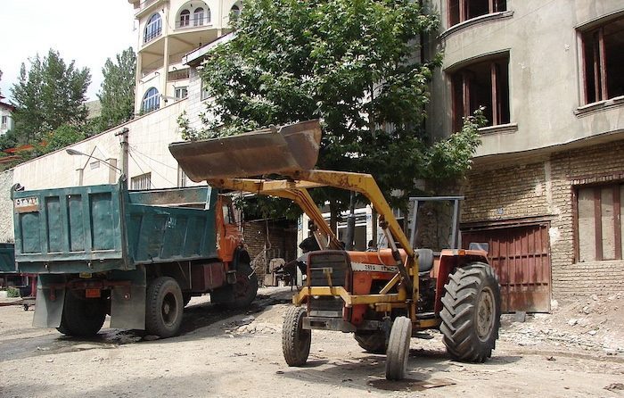 A green dump truck and a yellow and orange tractor in front of an apartment building on a dirt road.