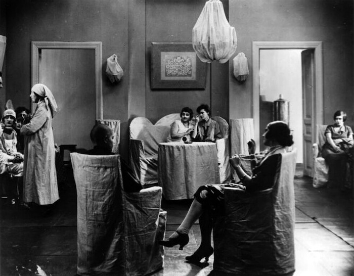 Seven actors sit in a living room full of furniture draped in white cloths.