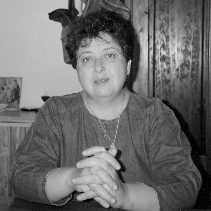 Black and white photograph of Regina Derieva, seated, looking at the camera with her hands folded on the table in front of her. 