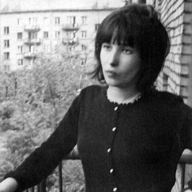 Elena Shvarts, with a dark bob and wearing a black cardigan, stands on a balcony of an apartment building.