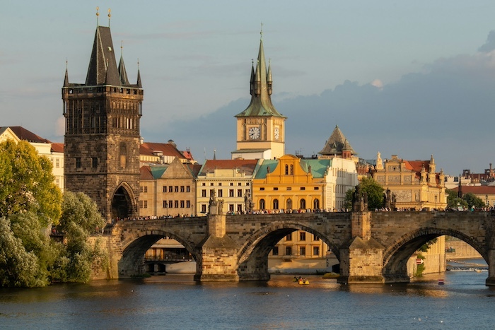 A stone bridge across a river. Behind it are a row of old buildings and a clock tower. 