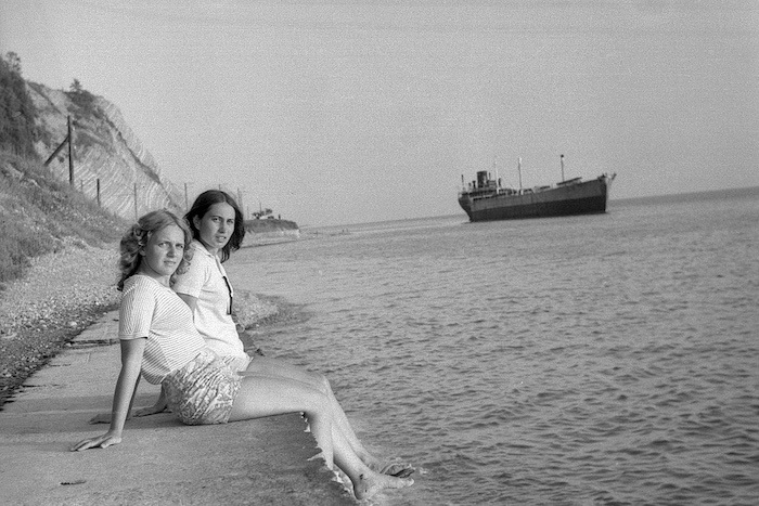 Two young women in short sit at a ledge on the ocean, their feet dangling in the water. A large ship sits in the background. 