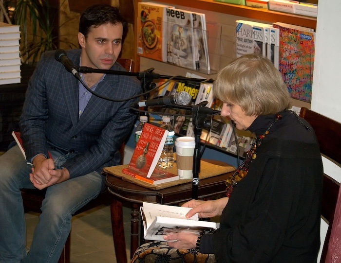 Keith Gessen sits behind a microphone and looks at Ludmilla Petrushevskaya, who sits next to him holding an open book.