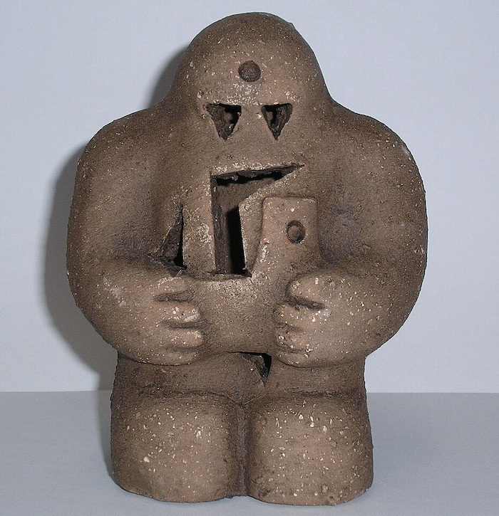 A small brown clay statue with a humanlike shape and a carved head and torso. 