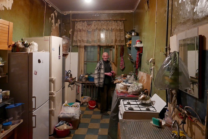 A bald man in a large sweater stands at the back of a cluttered apartment kitchen with peeling green walls and checkered linoleum tiles. 