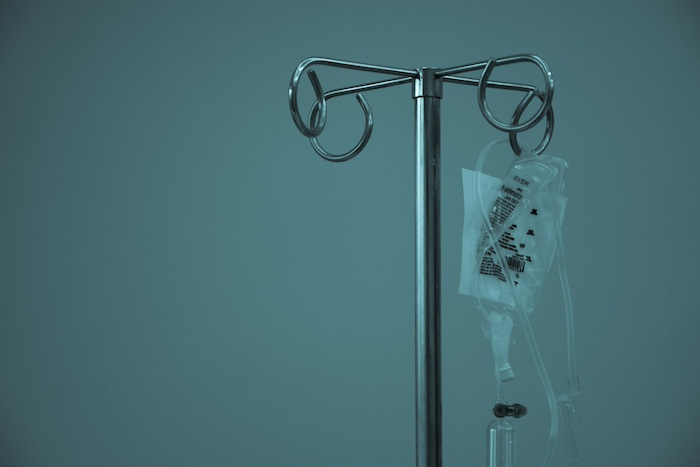 An IV drip hanging from a metal stand against a blue-grey background.