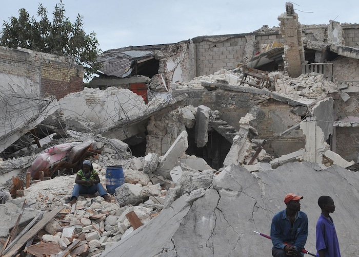 Three people sit on the rubble of a collapsed concrete building.