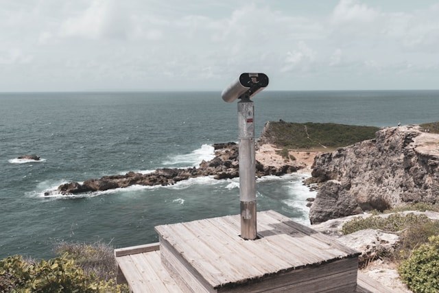 Image of binoculars mounted on a metal frame on a wooden box, overlooking a scene of the sea and rocky crags jutting out into the waters.