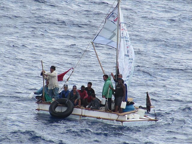 An image of 10 people aboard a small boat with a makeshift sail, in the waters.