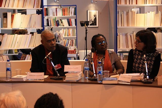 Author Évelyne Trouillot sitting at a conference table between two other speakers against a background of bookshelves.