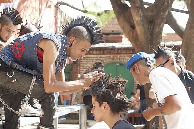 A man in a mohawk arranges another person's hair in a mohawk too, with people in the background wearing their hair in a range of hairstyles.