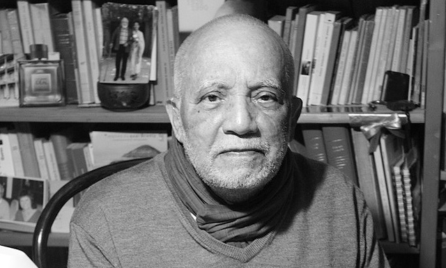 A black and white photo of Author René Depestre looking into the camera, sitting in a chair, against a backdrop of bookshelves.