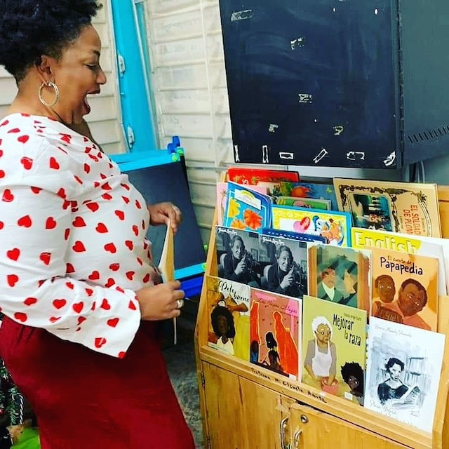 Author Yolanda Arroyo Pizarro smiling widely with excitement in front of a bookshelf with children's books.