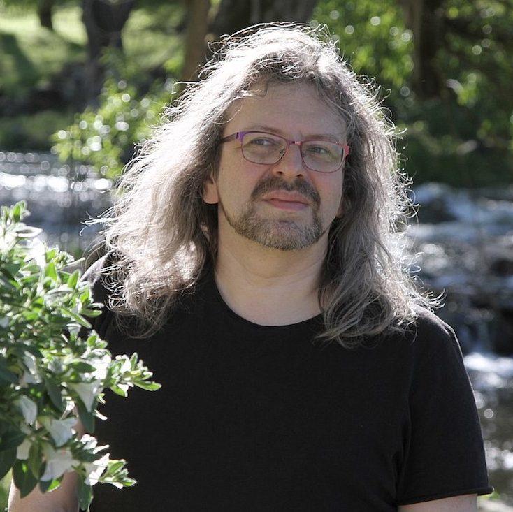 Portrait of author Dmitry Kuzmin with long grey hair, wearing a black t-shirt and glasses.