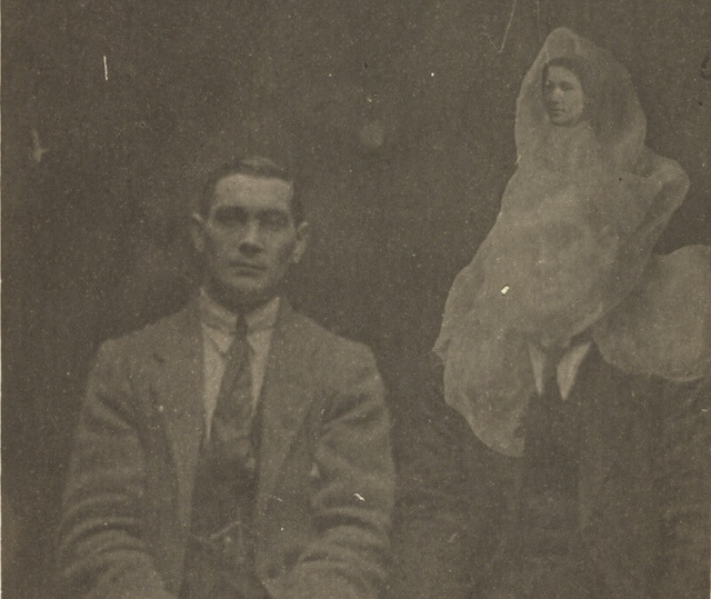 A sepia photograph of two men in suits staring at the camera. The ghost of a woman wrapped in a sheet is superimposed over one of the men.